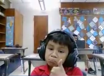 A young boy in a classroom. He is wearing headphones. He is giving a thumbs up while making a sad face.